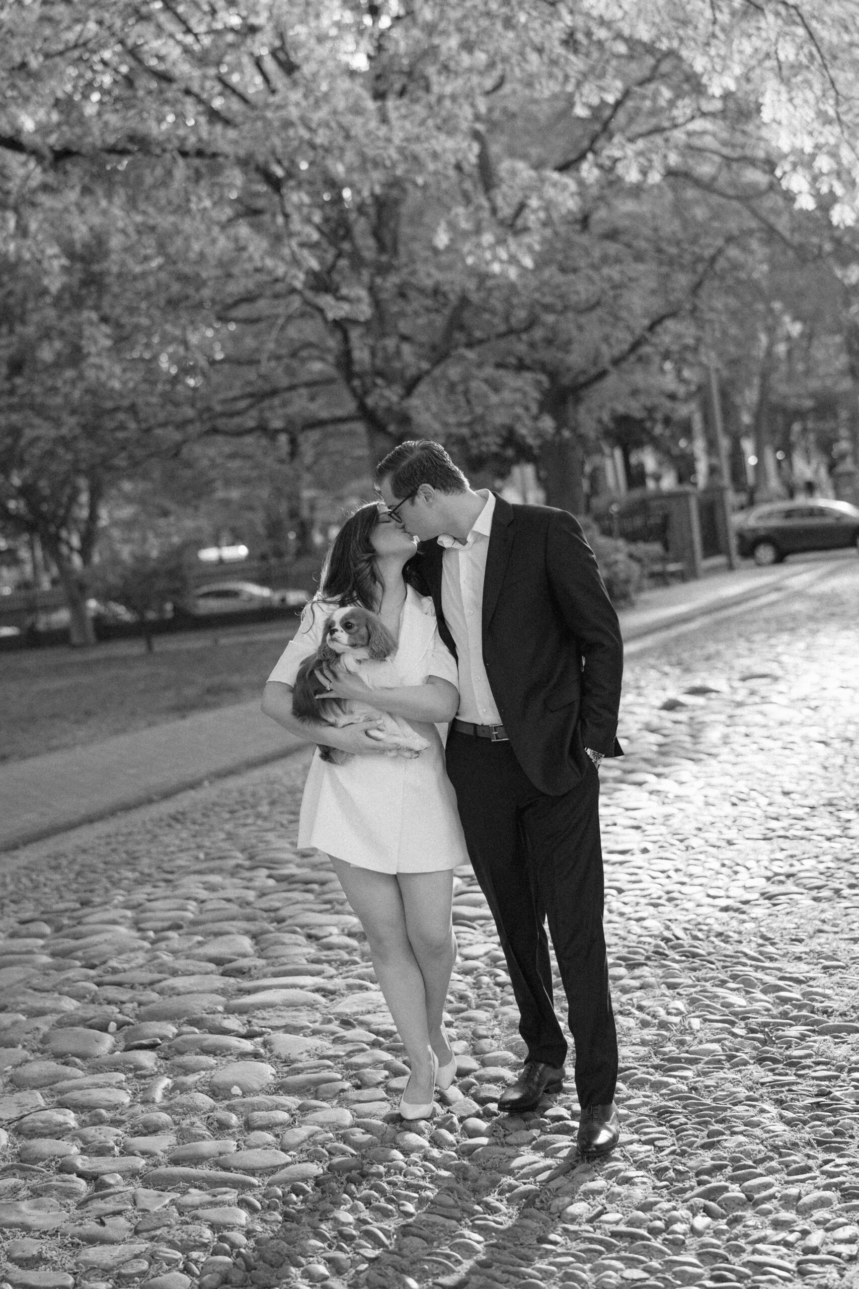 Black and white photo of couple kissing on the cobblestone streets of Old City Philadelphia. The bride-to-be is wearing a short, chic blazer dress and is holding her Cavalier King Charles Spaniel puppy.