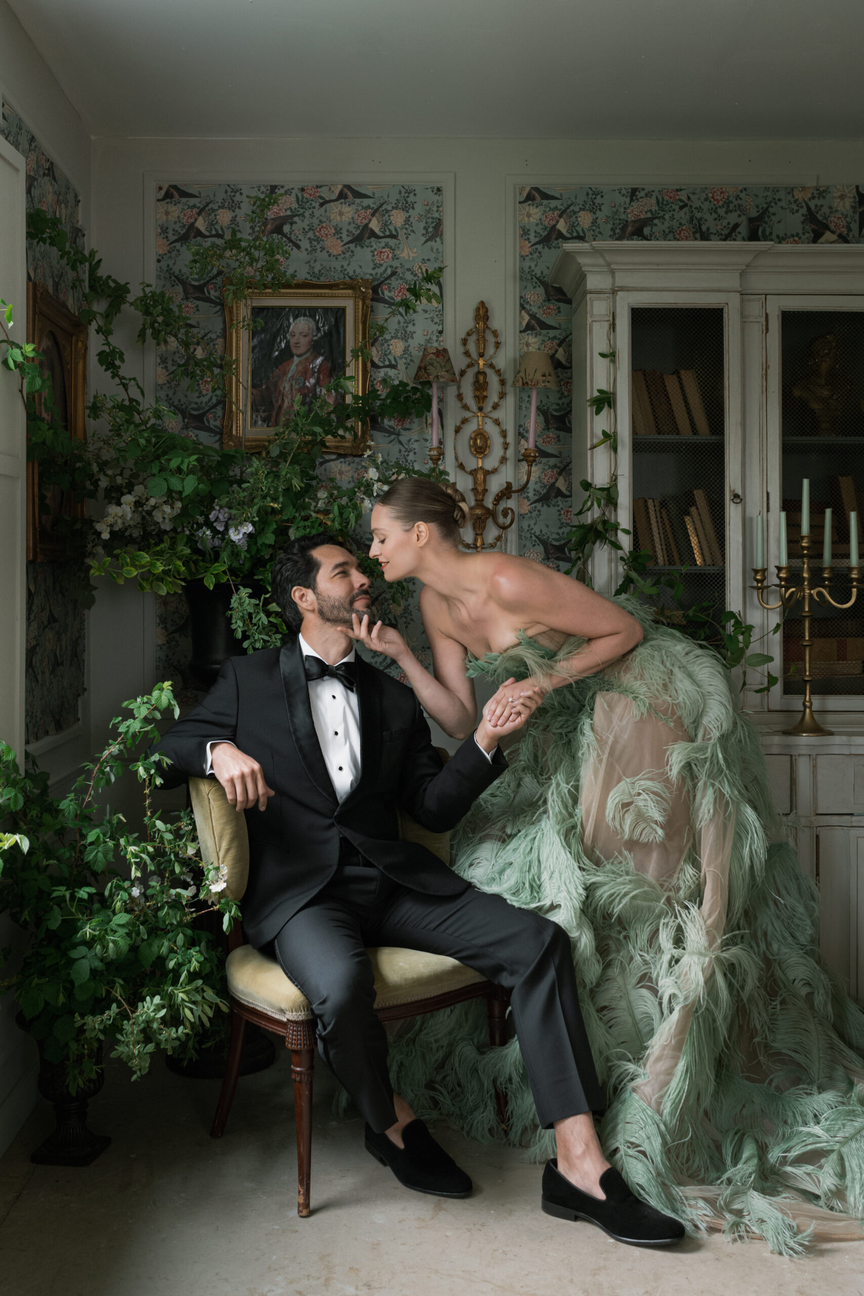 Bride in sage green feathered gown leans in to kiss chic groom in black tux. The room is decorative with wallpaper, organic florals, and antique furniture.