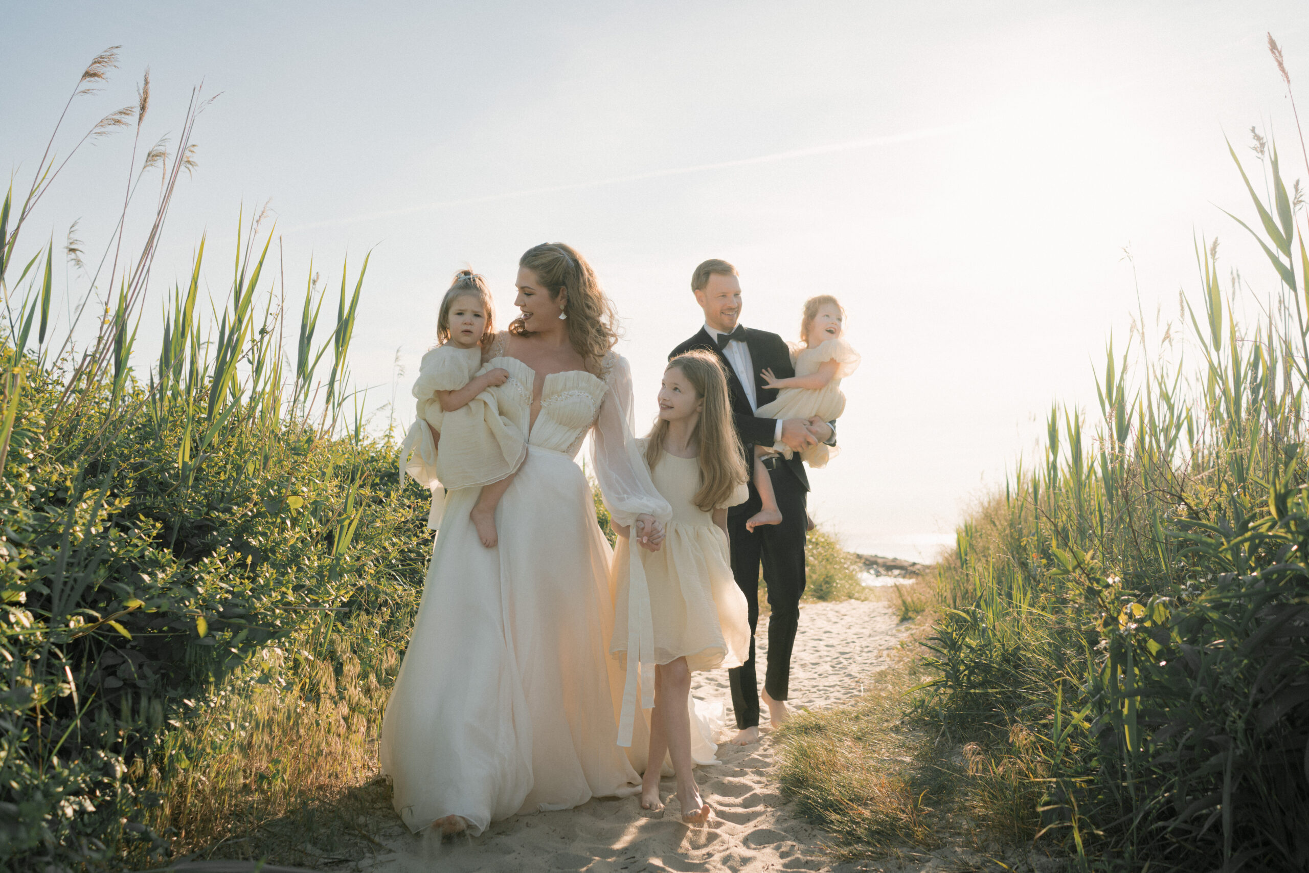 Family of five walks along a beachy pathway in Cape May surrounded by greens and sand with the sun shining behind them.
