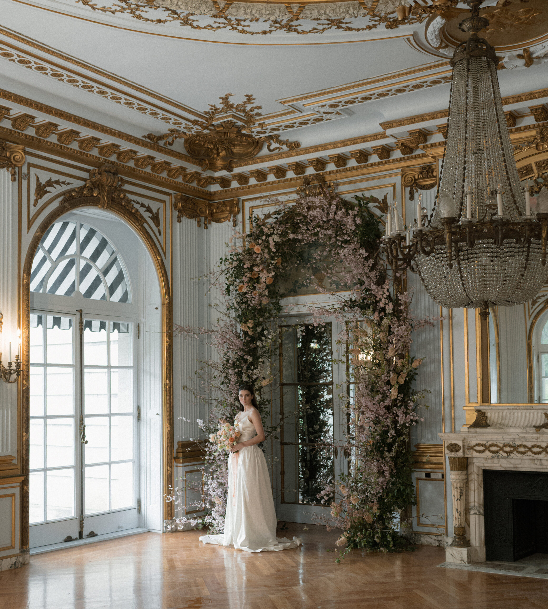 A large 14-foot floral arch lines a doorway in Elkins Estate, with an ethereal bride standing in the archway with a bouquet of flowers. The room is opulent and styled in Gilded Age grandeur.