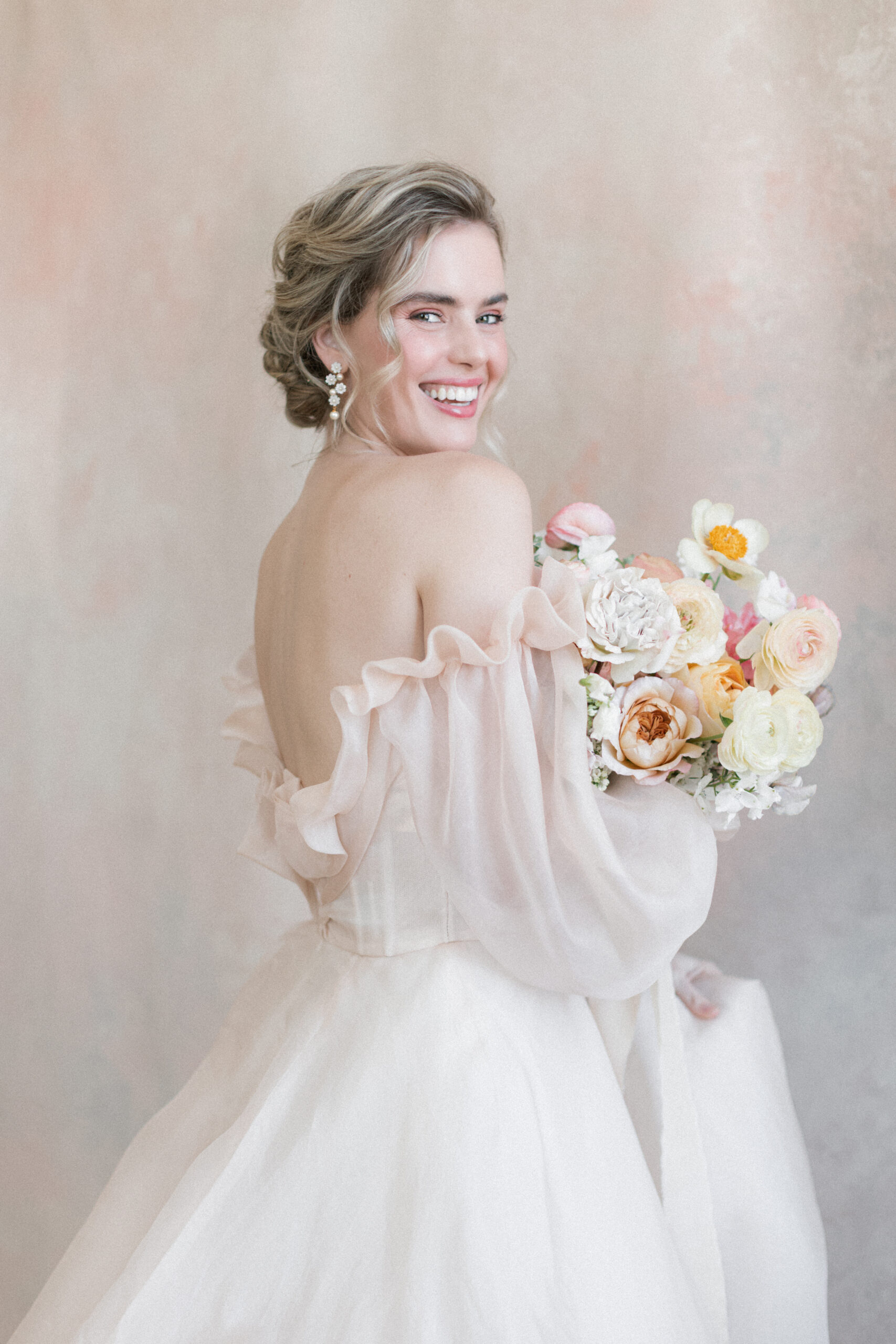 Bride in feminine Monique Lhuillier gown holds a bouquet with Claire de Lune peonies and mixed roses. She is standing in front of a soft blush painted backdrop.