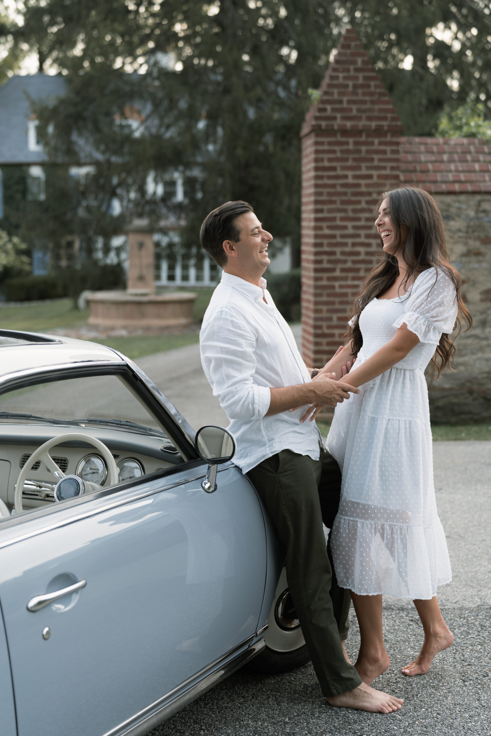 Couple playfully laughing alongside a dusty blue antique car at Willowbrook Farm in Chester County, PA. The setting feels European.