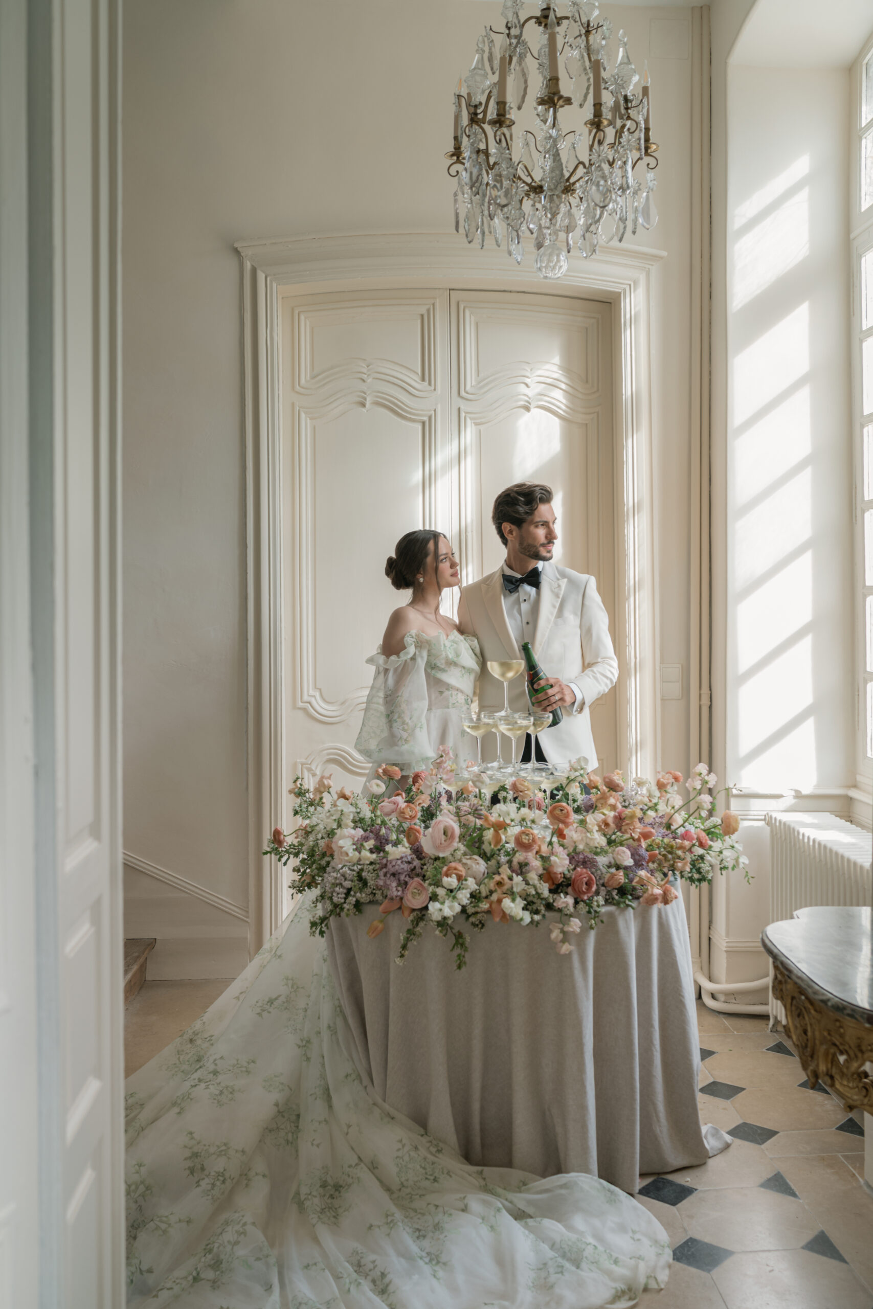 Bride and Groom stand aside a champagne tower surrounded by a floral meadow. They are in the sunlit hallway of a chateau in France.