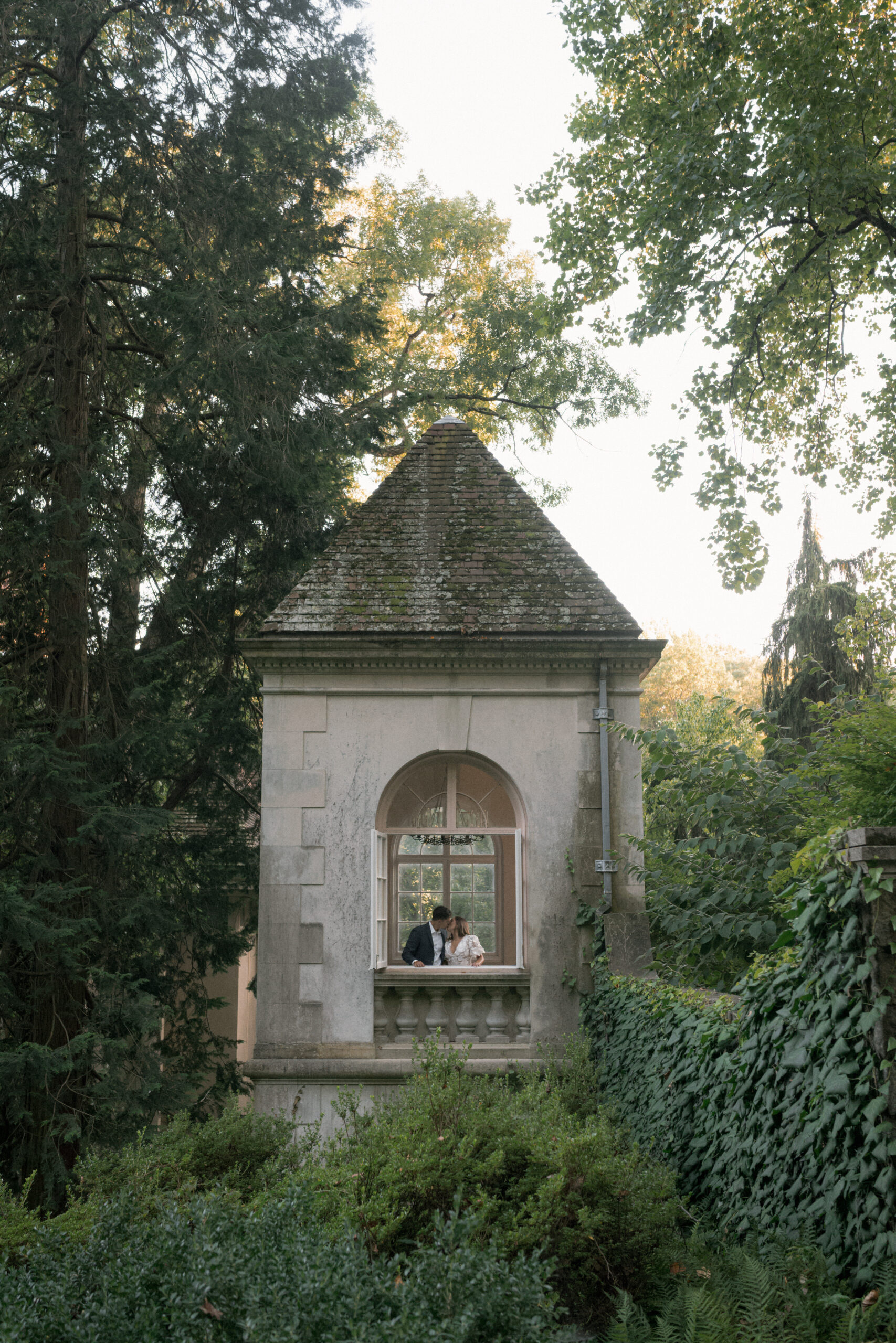 Couple shares a kiss inside an open window of Winterthur, surrounded by lush trees and the sunset.