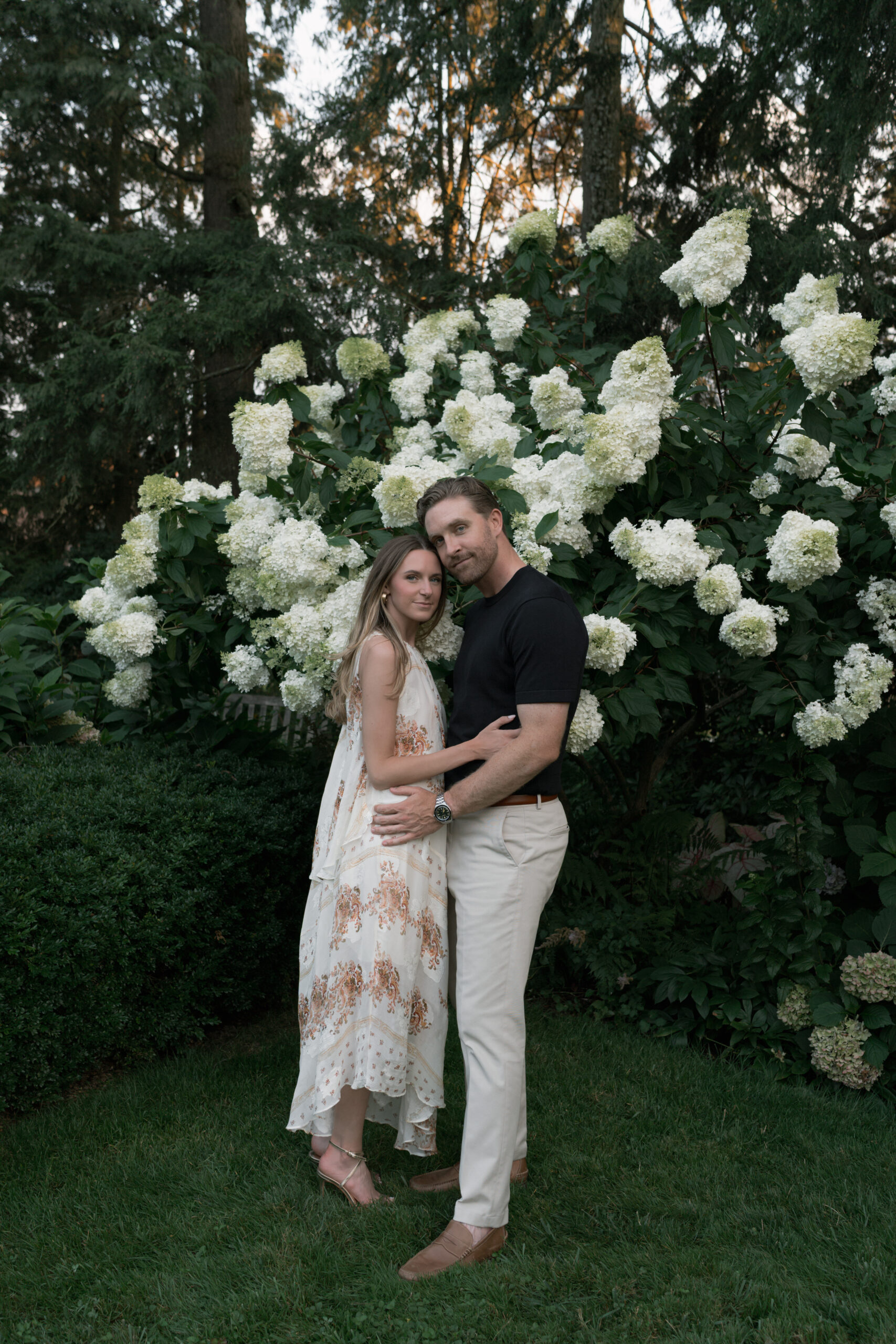Couple stands in front of hydrangea tree looking at the camera and embracing.