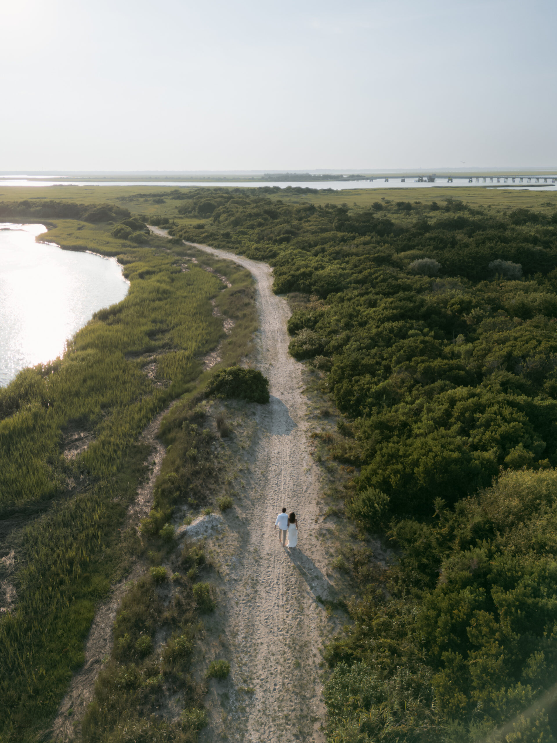 Drone shot of couple walking sandy beach path surrounded by grasses with the horizon in view.